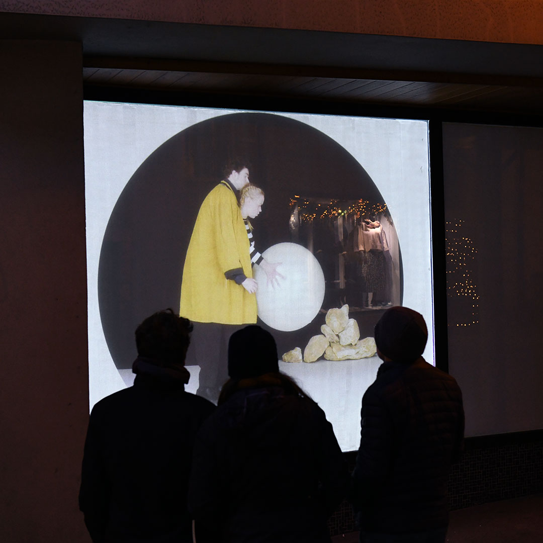 Trois passants regardent une projection vidéo projetée sur une vitrine à Porrentruy. La vidéo montre un duo de clowns jouant avec de grands ballons.
